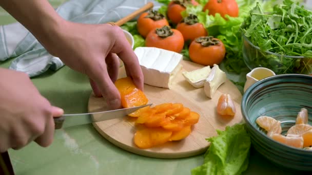 Preparación de ensalada vegana dietética a partir de caquis, mandarinas y lechuga. Vista desde arriba. movimiento de la cámara. Las manos humanas rebanan fruta para un desayuno o almuerzo saludable. — Vídeos de Stock