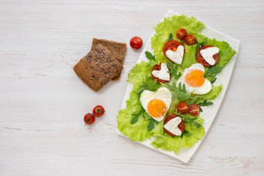 Sevgililer Günü 'nde kahvaltı. Bir tabak kalp şeklinde çırpılmış yumurta, domates, peynir, otlar ve tahıllı ekmek. Yukarıdan bak. Mesaj için yer var. Diyet kahvaltı..