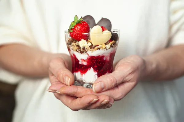 Kvinnors händer med ett glas söt krämig dessert på st. Alla hjärtans dag. dessert dekorerad med jordgubbar och choklad i form av ett hjärta — Stockfoto