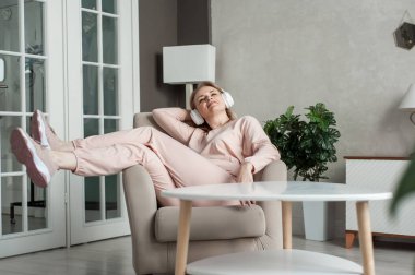 Beyaz kulaklıklı, ev elbiseli, orta yaşlı, güzel bir kadın bir sandalyeye oturup müzik dinler ve gülümser. Evde zihinsel sağlık, gençleşme ve rahatlama.