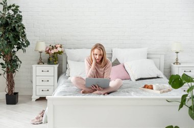 Evde takım elbiseli çekici, orta yaşlı sarışın bir kadın elinde dizüstü bilgisayarla yatakta oturuyor ve güçlü bir şaşkınlık yaşıyor. Çevrimiçi iletişim ve haberleri okumak. Metin için boşluk.