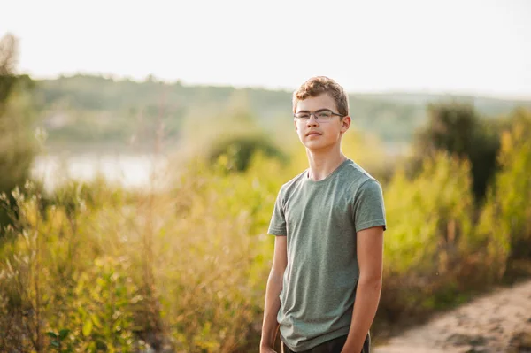 Portrait d'un adolescent en lunettes et un T-shirt vert sur fond de nature dans la lumière du coucher du soleil. — Photo