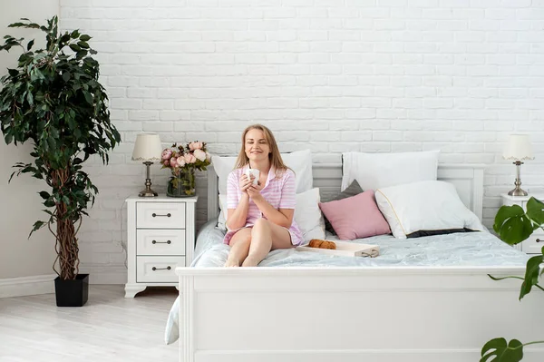 Atrakcyjna blondynka w paski w różowej piżamie siedzi na łóżku w sypialni wśród wewnętrznych roślin z filiżanką gorącego napoju w rękach i delektuje się spokojnym śniadaniem. Samoopieka. Obraz Stockowy