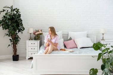 Pembe pijamalı çekici sarışın bir kadın yatak odasındaki bir yatağa oturur ve ev bitkilerinin arasında bir fincan sıcak içecek tutar ve pencereden dışarı bakar. Yatakta kahvaltı. Yavaş bir hayat. Dijital detoks