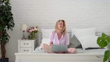 Apajamalı güzel, orta yaşlı bir kadın laptopuyla yatakta oturuyor. Çalış ve evden çalış. Çevrimiçi iletişim. Uzaktan eğitim. Metin için boşluk.