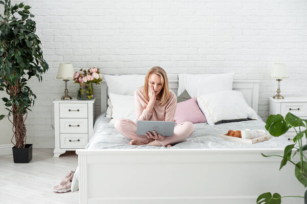 An attractive middle-aged blonde woman in a home suit sits on a bed with a laptop in her hands and expresses strong surprise. Online communication and reading the news. space for text.
