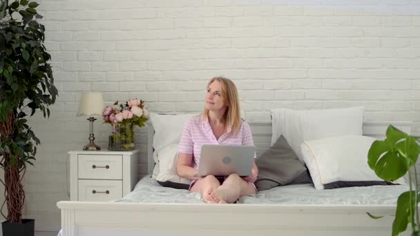 Glückliche blonde Frau mittleren Alters sitzt im Schlafanzug auf einem Bett mit Laptop im Schlafzimmer und drückt starke Überraschung und Freude aus. Ein unerwarteter Sieg oder eine gute Nachricht in Zeitlupe. — Stockvideo