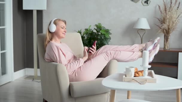 Hermosa mujer de mediana edad en ropa de casa se sienta en una silla con auriculares y un teléfono inteligente en sus manos. Aprendizaje y entretenimiento en línea. Descanso y recuperación en casa. Independiente. — Vídeo de stock