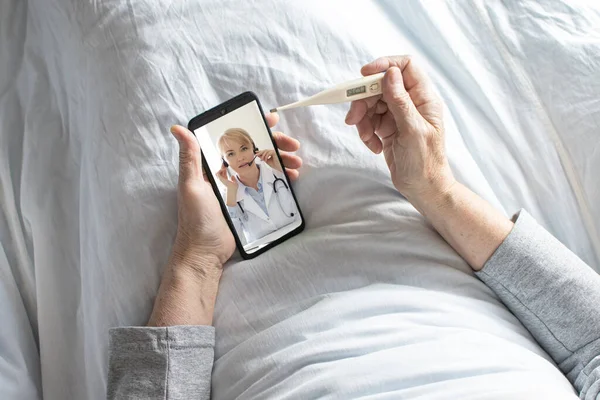 Starsza kobieta siedząca w łóżku pod białym kocem mierzy temperaturę za pomocą termometru elektronicznego podczas konsultacji online z lekarzemTelemedycyna Konsultacja online za pomocą smartfona. Zdjęcie Stockowe