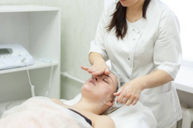 Yüz masajı. Kozmetik uzmanı çekici bir esmer kadın için yüz masajı yapıyor. Yüzün profesyonel bakımı ve kasları. Gençleştirici güzellik tedavileri.