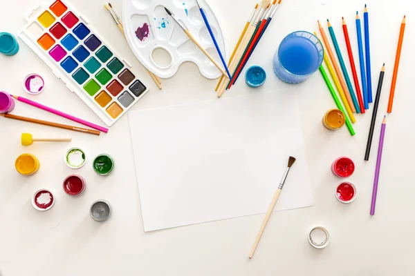 The artists workplace at the drawing lesson. A white sheet of paper surrounded by colorful paints and pencils. View from above. Hobby. multicolored background. A place for text or a picture. — Stock Photo, Image