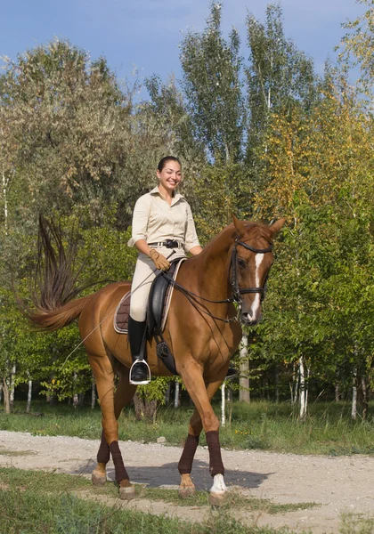 Beautiful smiling women riding a brown horse through woodland — Stock Photo, Image