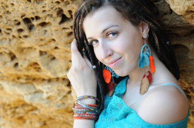 Sarı taş kayanın yanında poz veren, deri tüy küpeler takmış kameraya bakan mutlu gülümseyen genç kadın portresi