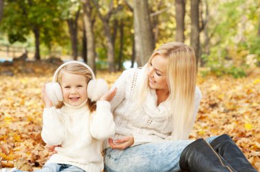 Happy joyful family portrait, blonde mother and blonde daughter outdoor in autumn park, girl dressed in fur earmuffs