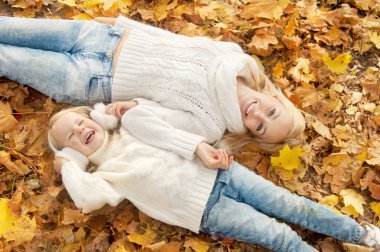 Mutlu aile anne ve kızı rahatlayıp sonbahar parkında akçaağaç yapraklarına uzanır.