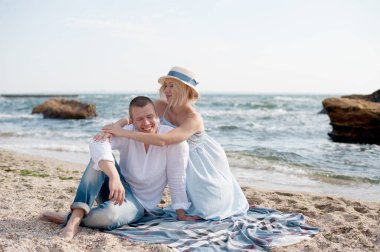 Happy future parents couple have a rest sitting near sea, husband hugs belly his pregnant wife