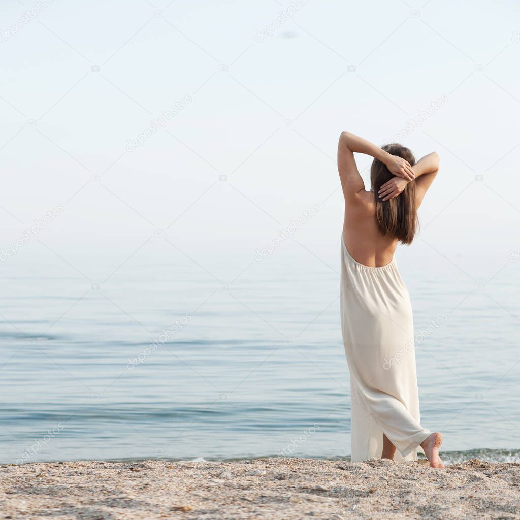 Youg calm dreaming woman standing alone near sea, looking on the water, back view hands up, outdoor