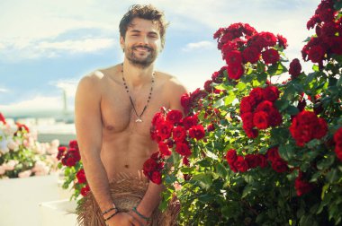 Gül çalılarının yanında duran yakışıklı, çıplak, gülümseyen genç adamın portresi.