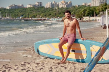 Genç sörfçü üstsüz açık hava portresi sörf tahtasında oturuyor.