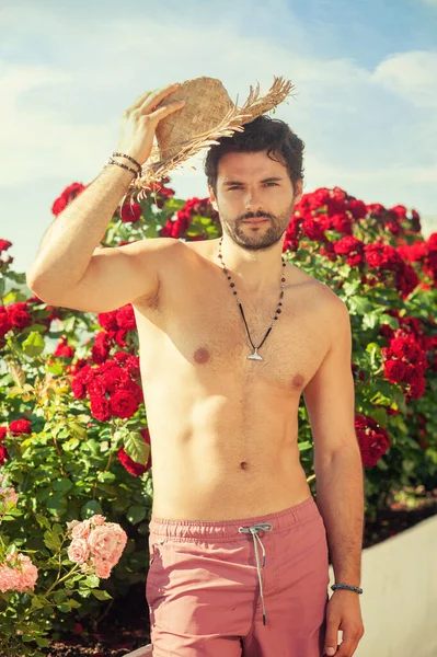 ハンサムな裸の男の肖像近くに立っているバラの茂みとともにわら帽子 — ストック写真