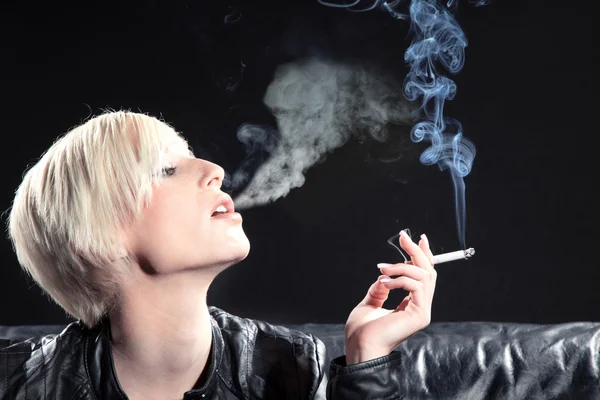 Blonde girl smoking a cigarette on a black couch — Stock Photo, Image