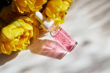 Şeffaf cam bir şişede çivi ve tırnak serumu. Beyaz arka planda güzel sarı, duble şakayık laleleri var. 