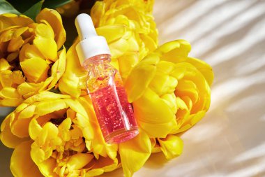 Şeffaf cam bir şişede çivi ve tırnak serumu. Beyaz arka planda güzel sarı, duble şakayık laleleri var. 