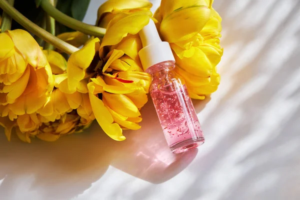 Şeffaf cam bir şişede çivi ve tırnak serumu. Beyaz arka planda güzel sarı, duble şakayık laleleri var. 