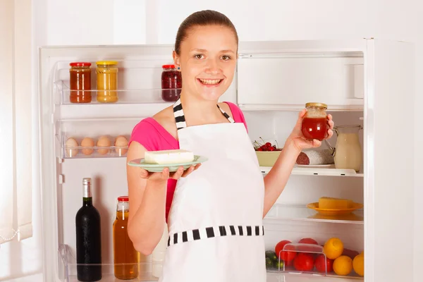 Une jeune femme avec de la confiture et du beurre à la main devant le réfrigérateur ouvert. Nourriture, lait, vin rouge et jus en arrière-plan . — Photo