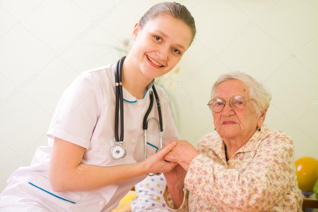 A young doctor, nurse visiting an elderly sick woman holding her hands with caring attitude.