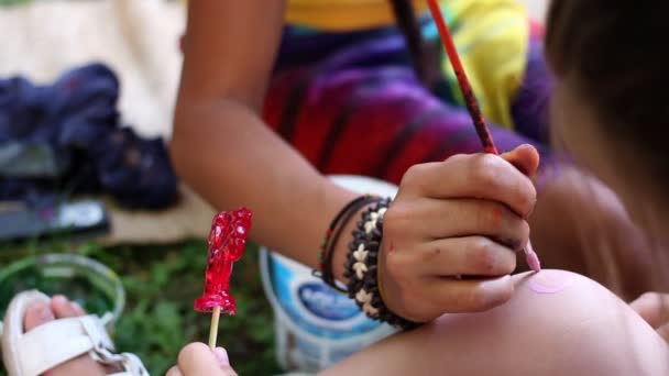 A cute little European girl sits patiently while the artist paints a pink spiral on her knee. — Stock Video