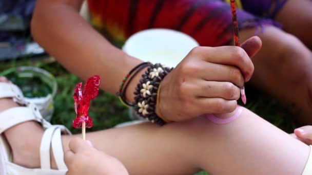 A cute little European girl sits patiently while the artist paints a pink spiral on her knee. — Stock Video