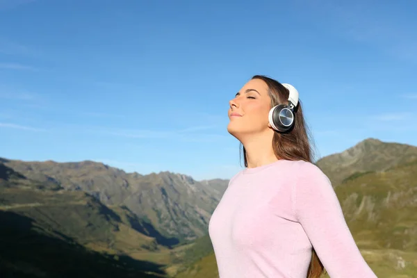 Relaxed woman wearing wireless headphones listening audio guide breathing fresh air in the mountain