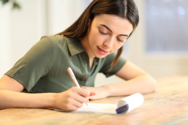 Mutlu kadın evdeki masadaki kağıt deftere notlar alıyor.