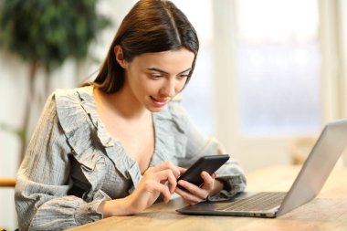 Evde dizüstü bilgisayarın yanında cep telefonunu kontrol eden tatmin olmuş bir kadın.