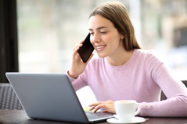 Kahve dükkanında dizüstü bilgisayar kullanan mutlu bir kadın akıllı telefondan konuşuyor.