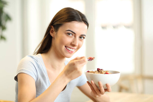 Happy woman eating cereals from a bowl holding spoon looking at you in a house