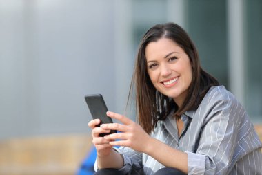 Happy student holding smart phone looks at you in a campus