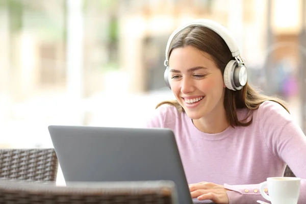Happy woman with wireless headphones laughing checking laptop in a coffee shop