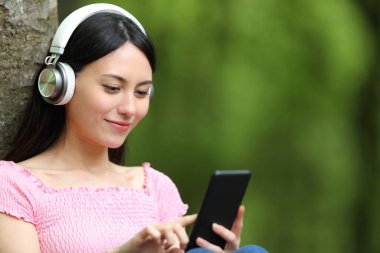 Asyalı kadın müzik dinliyor. Parkta oturmuş akıllı telefonları kontrol ediyor.