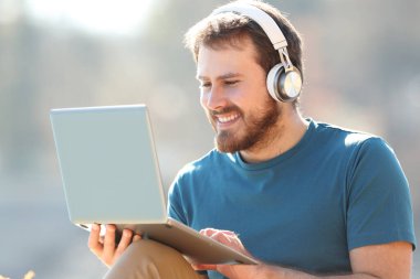 Dışarıda dizüstü bilgisayar kullanan, kablosuz kulaklık takan mutlu adam.