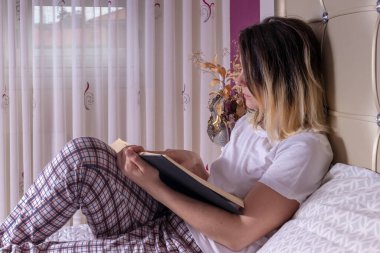 Kadın yatakta oturmuş beyaz tişört ve ekose pijamayla kitap okuyor.