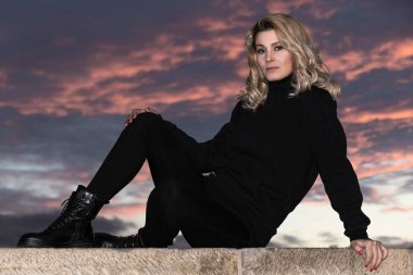 Gün batımında siyah giyinmiş, duvarda oturan sarışın kadın portresi.