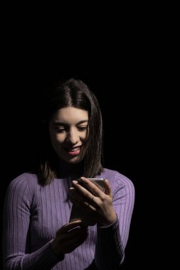 Gülümseyen esmer kadın siyah arka planlı cep telefonuna bakıyor.