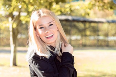 Güzel sarışın kadın, parkta siyah giyinmiş mutlu bir şekilde gülümsüyor.