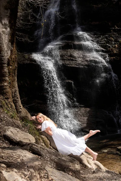 白いドレスを着たブロンドの女性が滝の後ろに木の上に横たわっていた — ストック写真