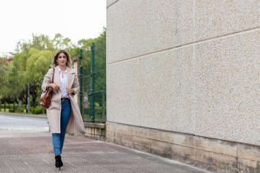 Sarışın bir kadın sokakta çalışıyor, bej bir ceket, beyaz gömlek ve kot pantolon giyiyor.