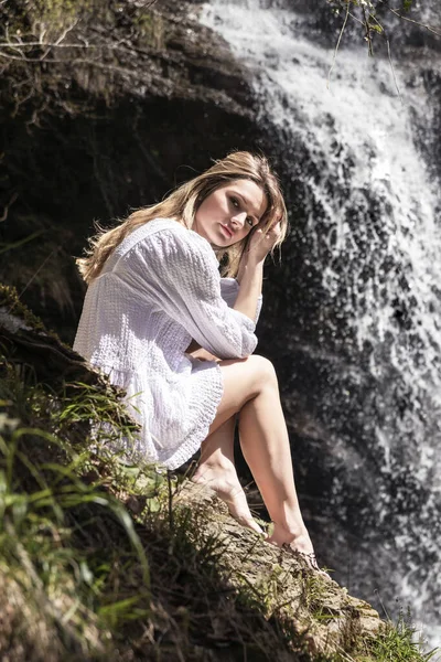 滝の前の森に座っている白いドレスのブロンドの女性 — ストック写真