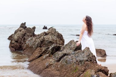 Genç esmer kadın plajda beyaz elbiseli bir kayaya yaslanıyor.