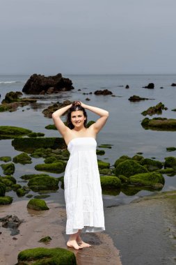 Beyaz elbiseli genç esmer kadın sahilde poz veriyor.